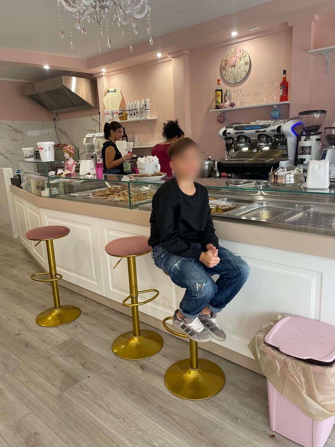 Interno di una caffetteria con bancone in vetro, sgabelli dorati e arredi rosa pastello.