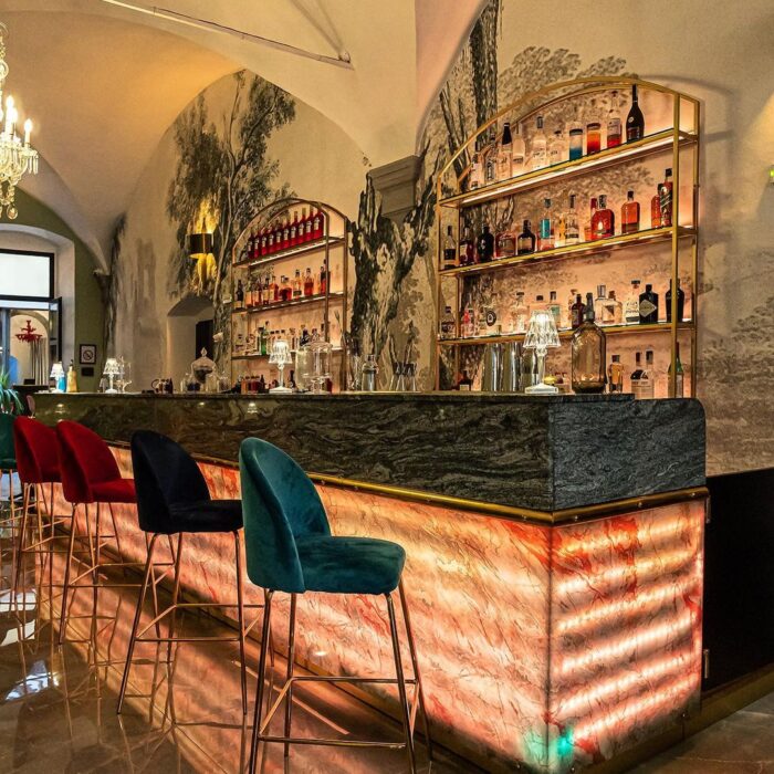 Bar Elegante con Bancone in Marmo Retroilluminato - Soluzioni di Design di Agape Arredo Bar Interno di un bar elegante con bancone in marmo retroilluminato, scaffali dorati e sgabelli colorati.