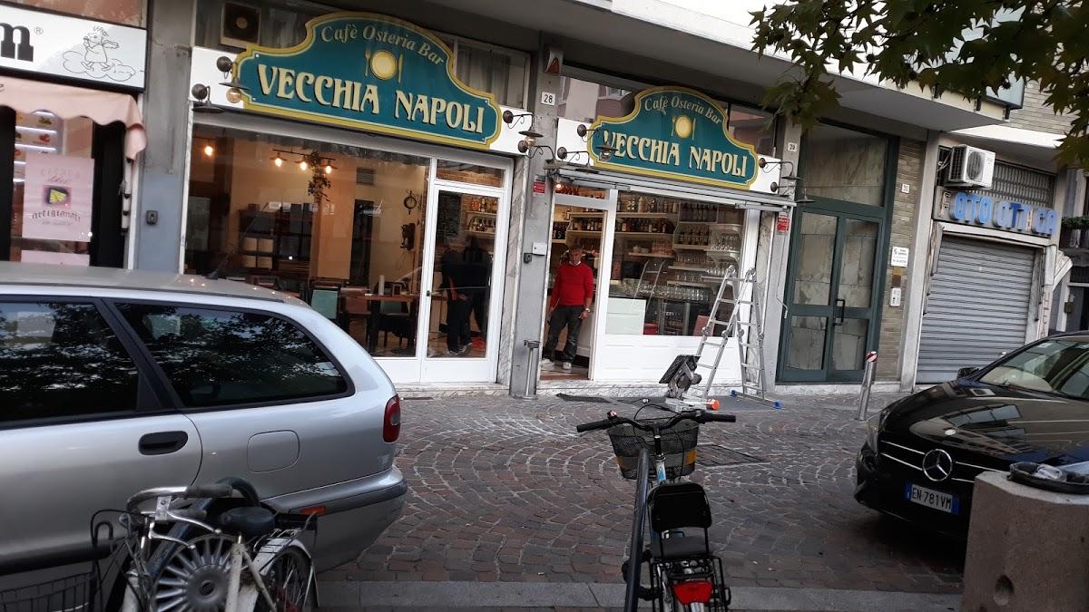 Facciata del bar Vecchia Napoli con due ingressi affiancati, insegne verdi e gialle, e lavoratori visibili all'interno mentre effettuano preparazioni. Auto e biciclette sono parcheggiate davanti al locale