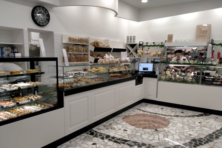Interno di un negozio di alimentari con banconi espositivi pieni di dolci, pane, salumi e formaggi. Il negozio ha un pavimento in mosaico e un ambiente ben illuminato e accogliente.