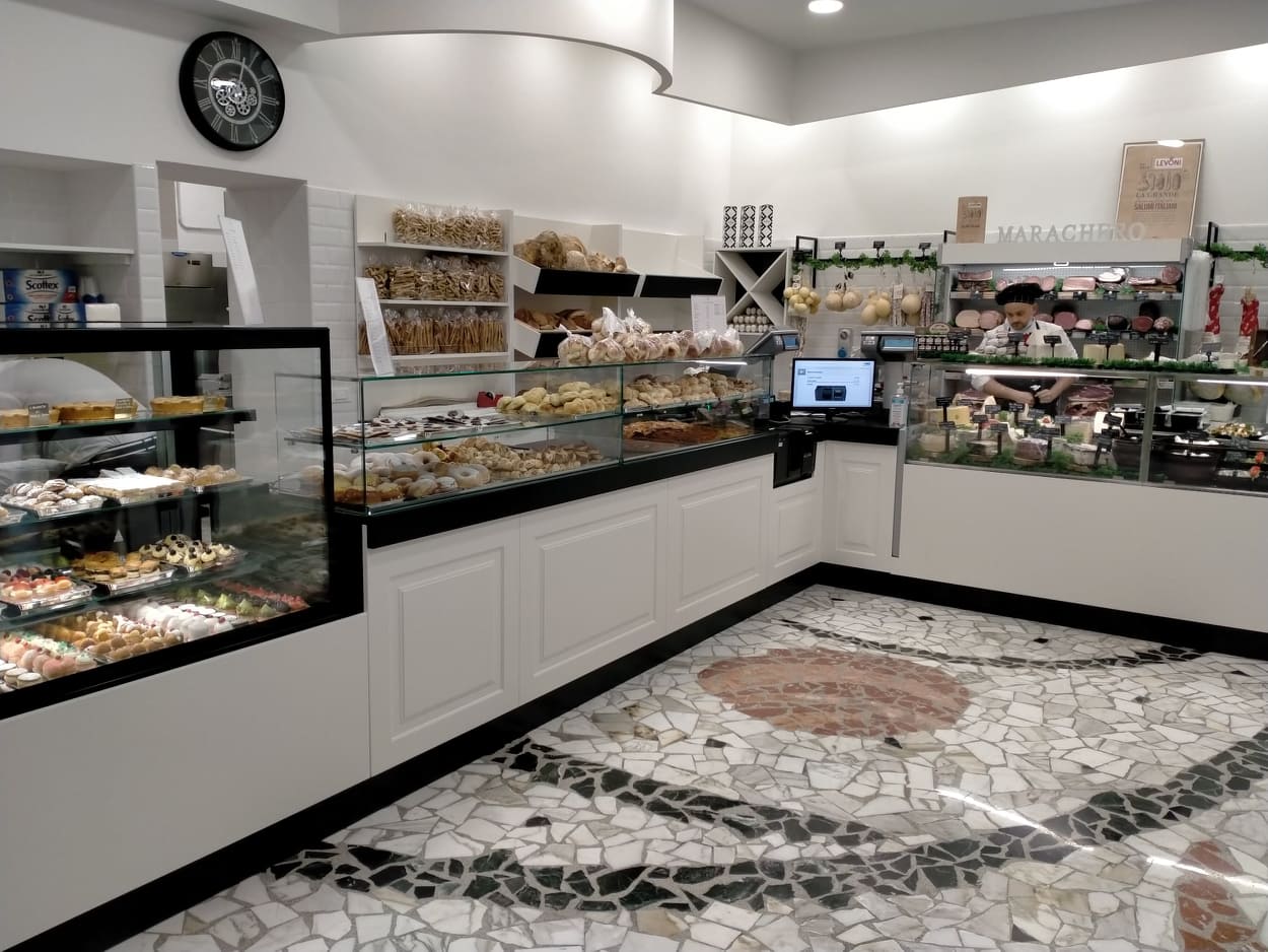 Interno di un negozio di alimentari con banconi espositivi pieni di dolci, pane, salumi e formaggi. Il negozio ha un pavimento in mosaico e un ambiente ben illuminato e accogliente.