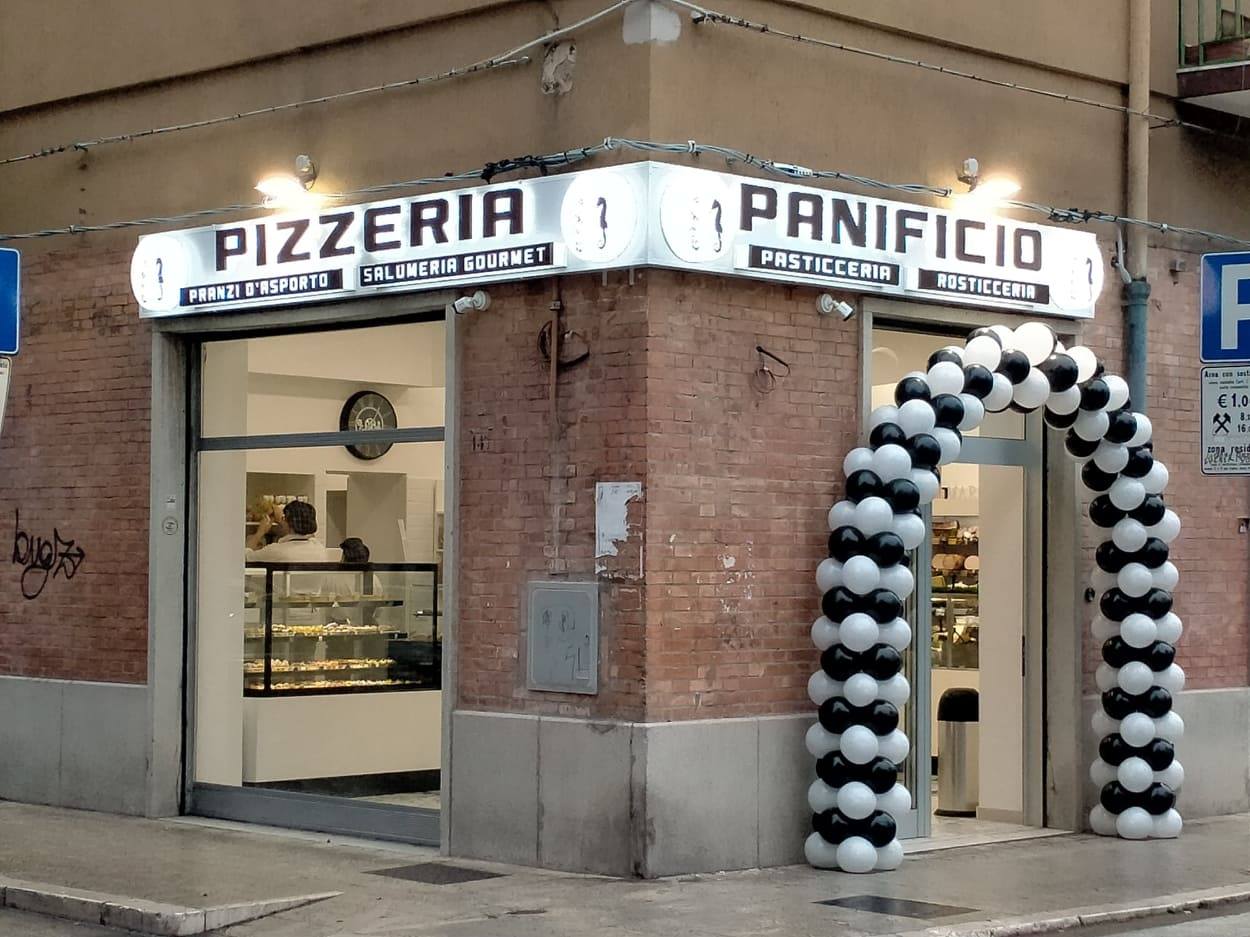 Facciata di una pizzeria e panificio con insegne luminose e un arco di palloncini bianchi e neri all'ingresso. Il negozio offre pranzi d'asporto, salumeria gourmet, pasticceria e rosticceria.