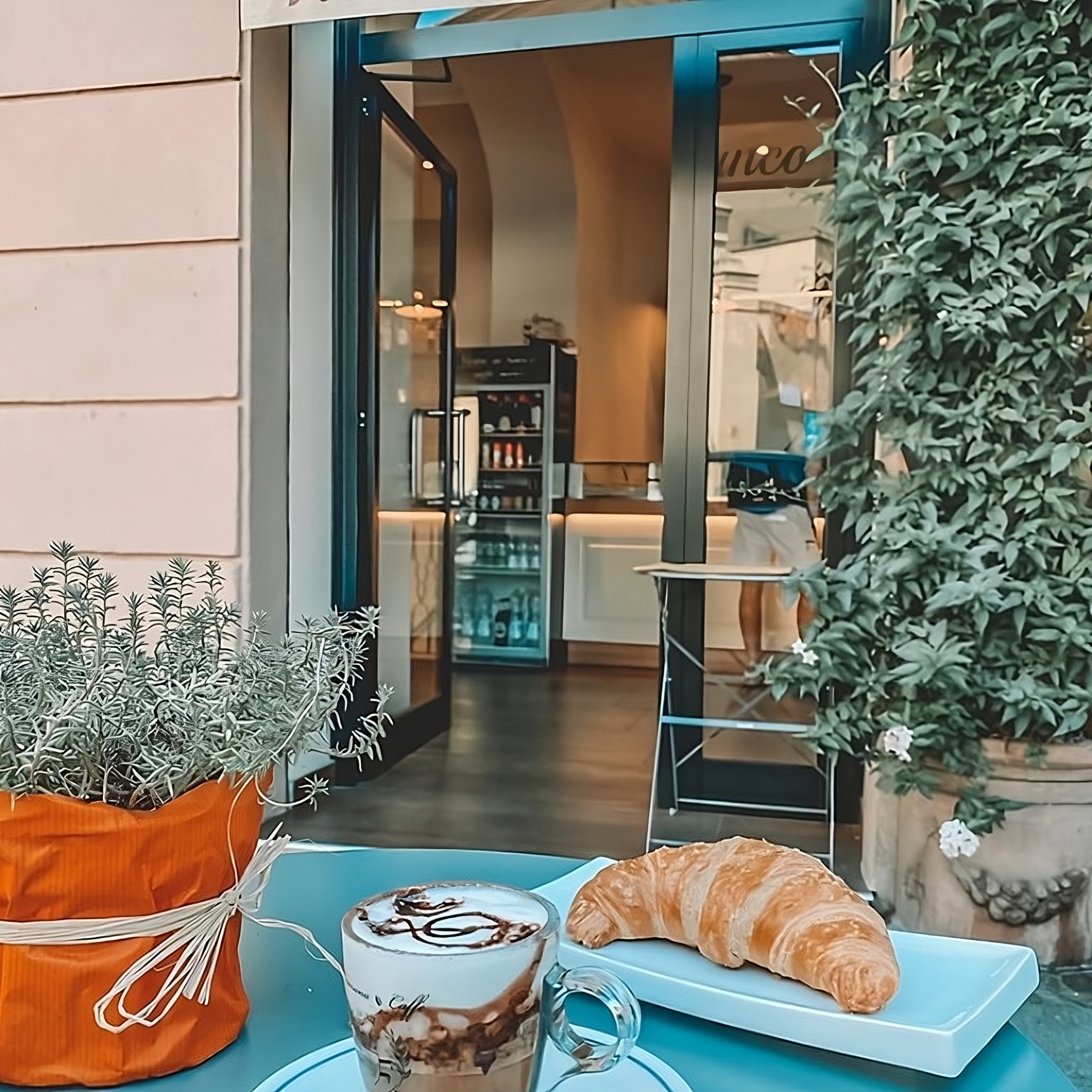 Tavolino con cornetto e cappuccino, ingresso del locale e bancone laccato bianco crema e legno venato, Roma