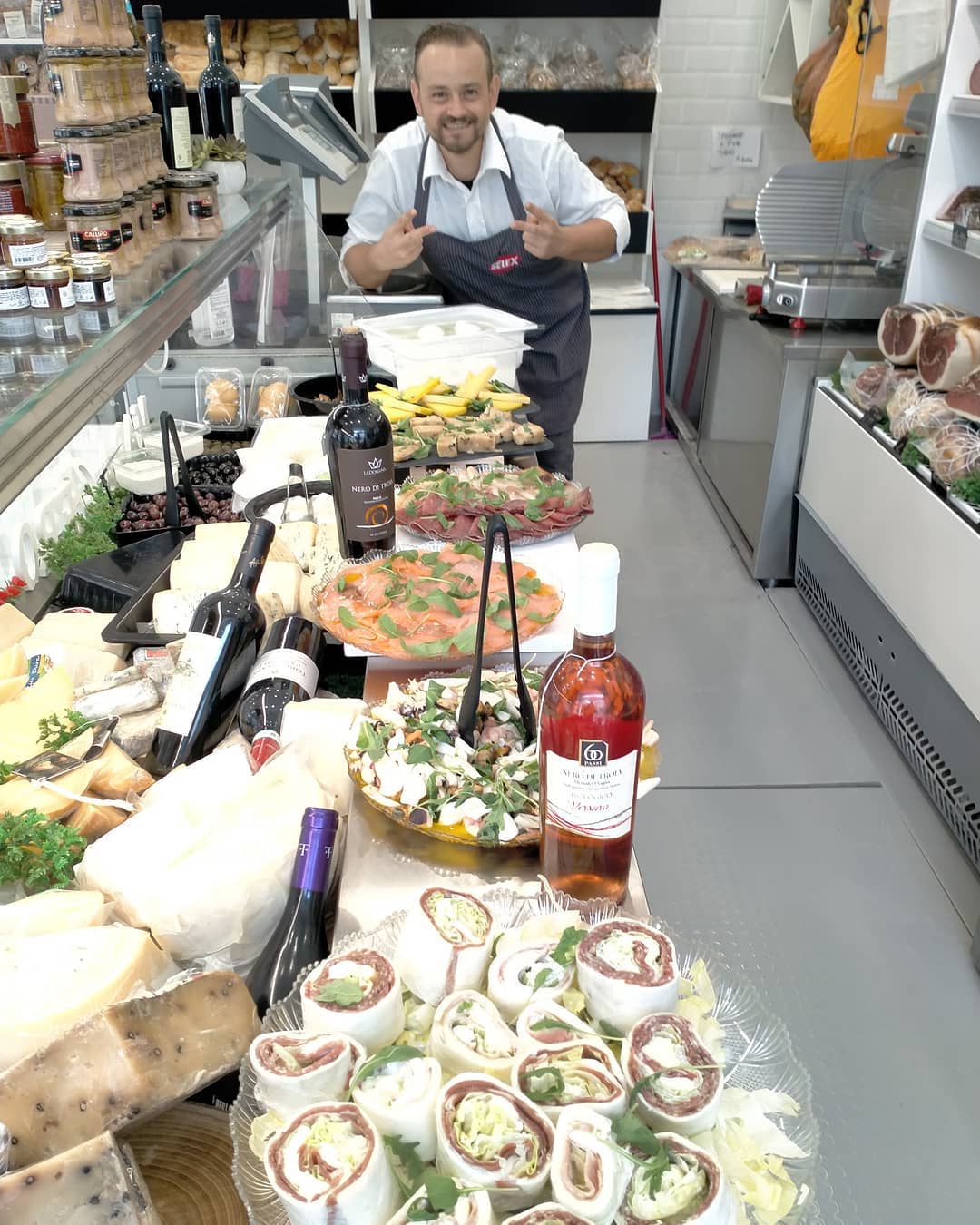 Negozio di alimentari con bancone pieno di cibo fresco, formaggi, salumi, vini e insalate. Un negoziante sorridente dietro il bancone.