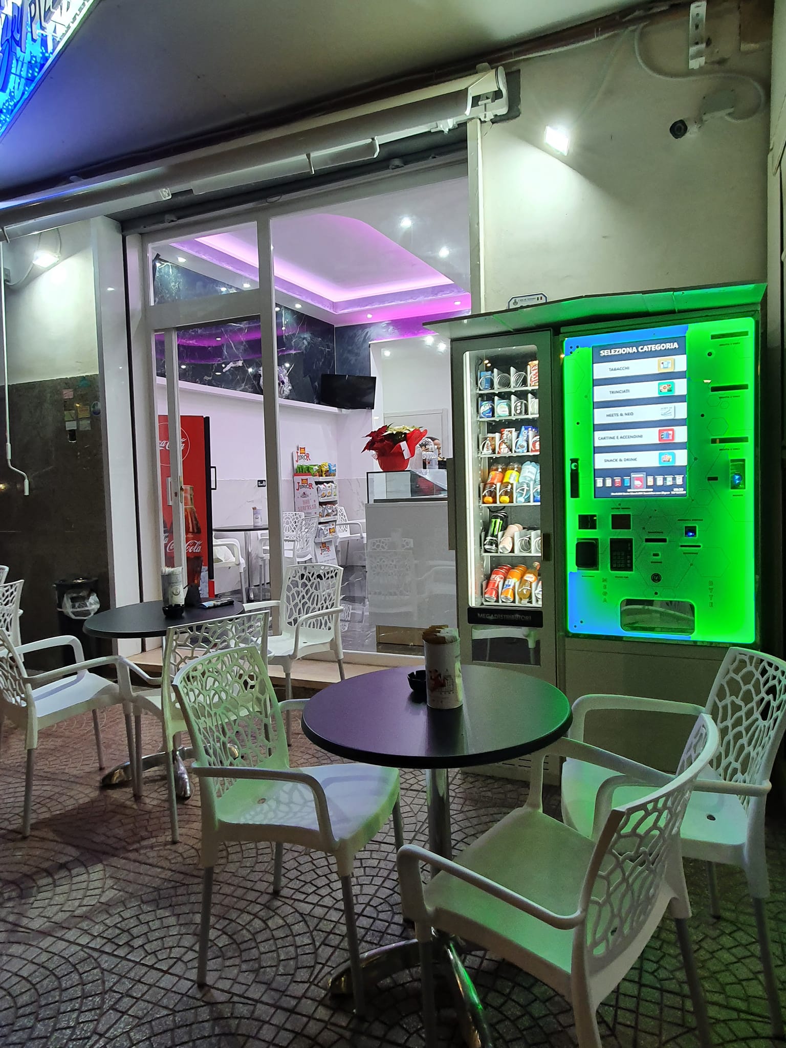 Esterno di un locale con tavoli e sedie bianche, distributore automatico e illuminazione moderna