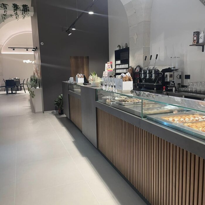 Interno di un locale moderno con bancone in legno e vetro, illuminazione a soffitto, e pavimento chiaro