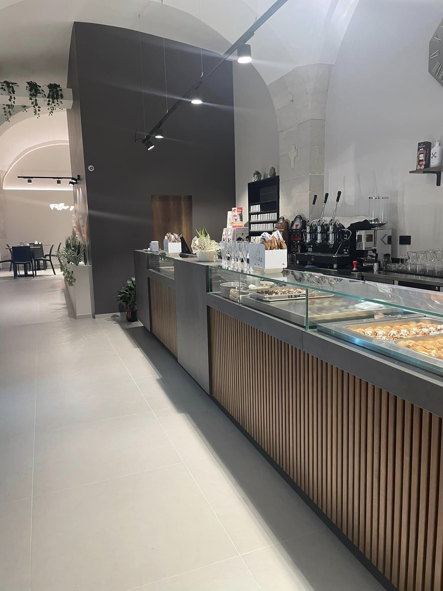 Interno di un locale moderno con bancone in legno e vetro, illuminazione a soffitto, e pavimento chiaro