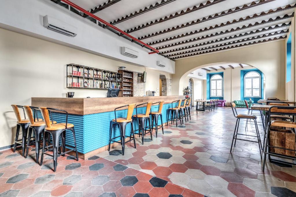 Bar moderno e accogliente con un banco bar in legno e dettagli blu, sedie alte in legno e metallo, scaffalature con bottiglie di liquore, pavimento a piastrelle esagonali e soffitto decorativo con travi a vista.