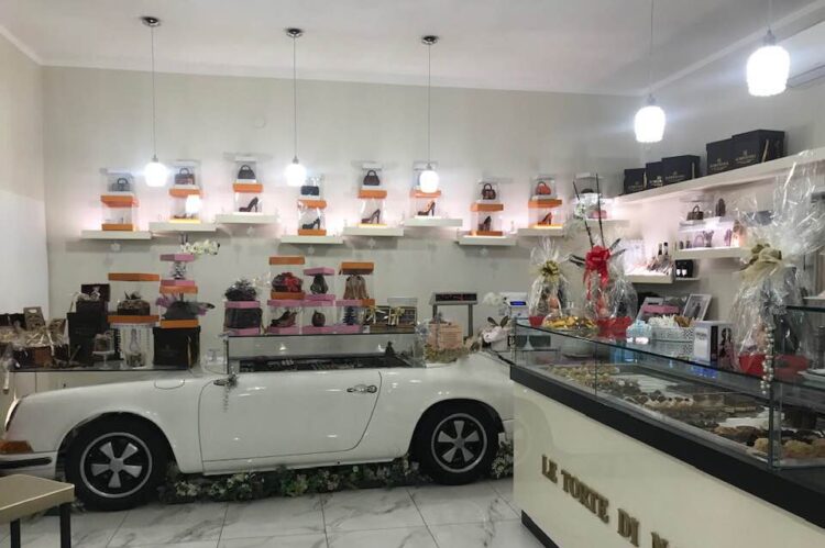 Interno del locale con bancone a forma di auto bianca, espositori con borse e scarpe, e vetrina pasticceria
