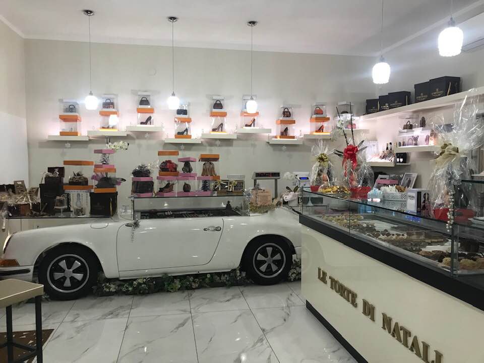 Interno del locale con bancone a forma di auto bianca, espositori con borse e scarpe, e vetrina pasticceria