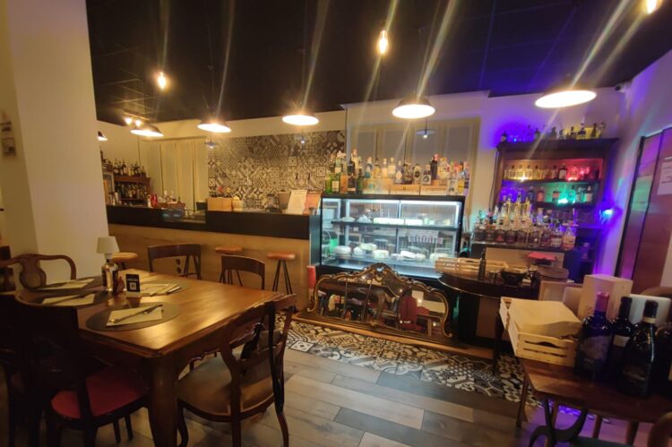Bancone bar lato cliente con vetrina cubica da 150 cm, sgabelli, lampade sconce industrial, tavoli in legno massello e sedie, realizzato da Agape Arredo Bar