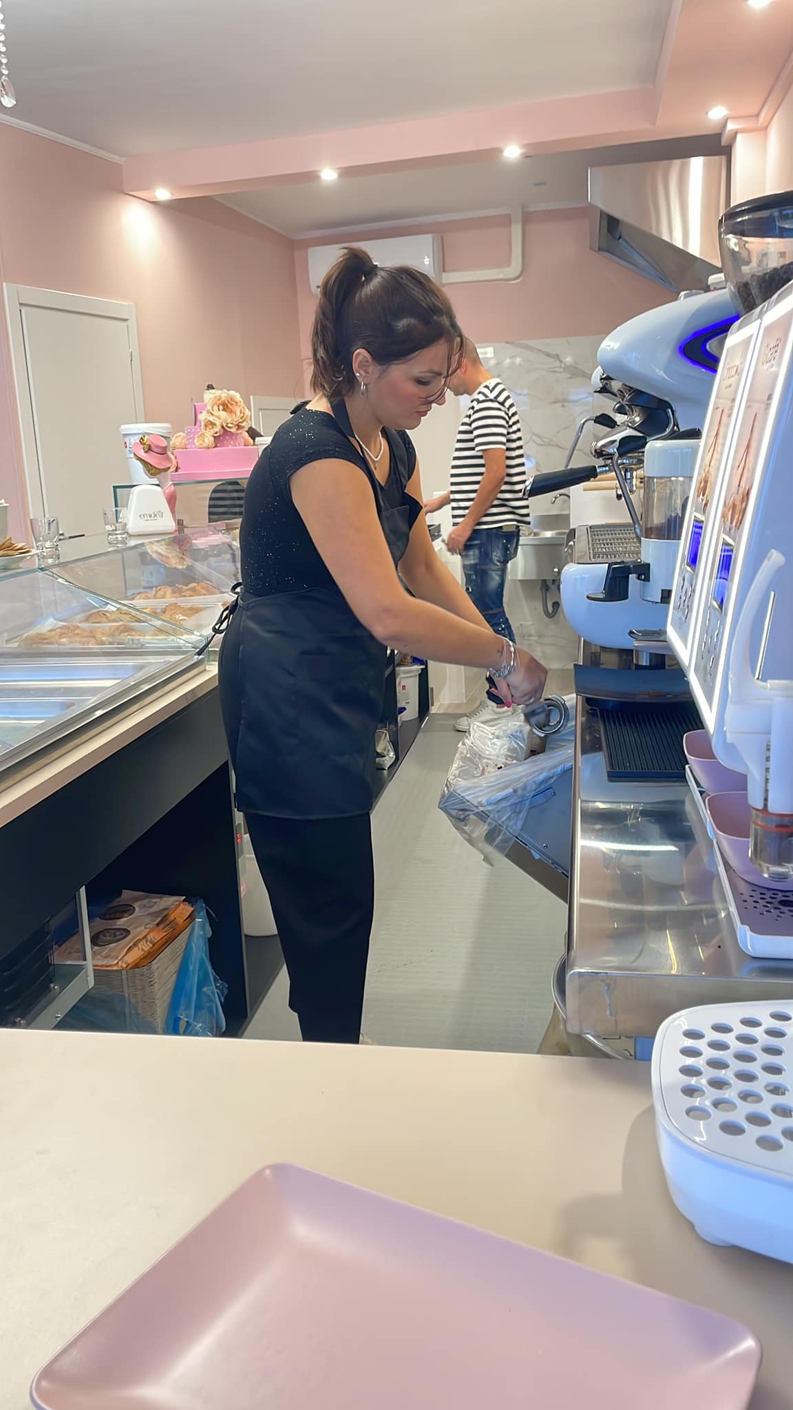 Interno di una caffetteria moderna con una barista al lavoro dietro un bancone di vetro. Pareti rosa e un ambiente accogliente. Realizzato da Agape Arredo Bar