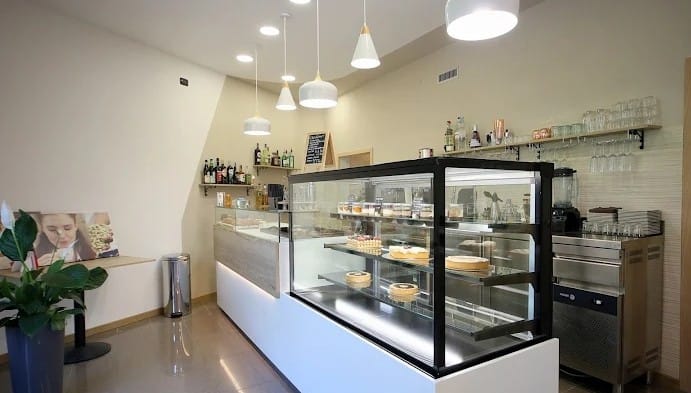 Interno di un bar pasticceria moderno con una vetrina di dolci, un bancone in legno e illuminazione pendente. Realizzato da Agape Arredo Bar.