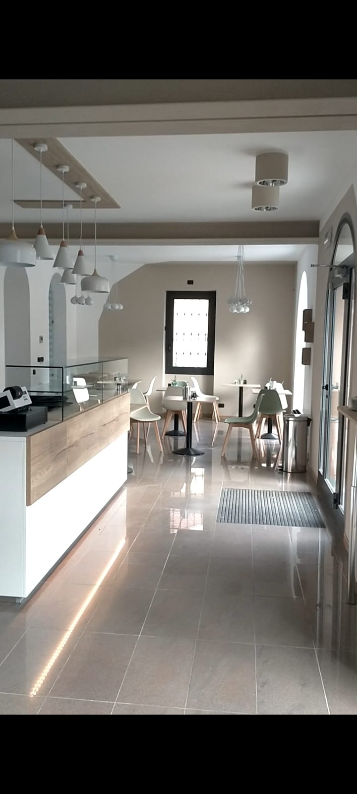 Interno di un bar moderno con un bancone in legno, sedie bianche, tavoli neri e illuminazione pendente. Realizzato da Agape Arredo Bar.