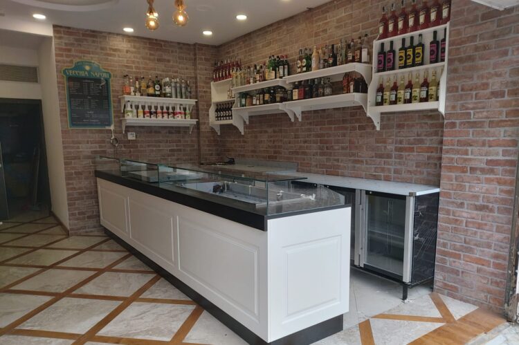Interno di un bar moderno con un bancone in legno bianco, ripiani con bottiglie di liquori e una parete di mattoni a vista. Il pavimento &egrave; in marmo con intarsi in legno.