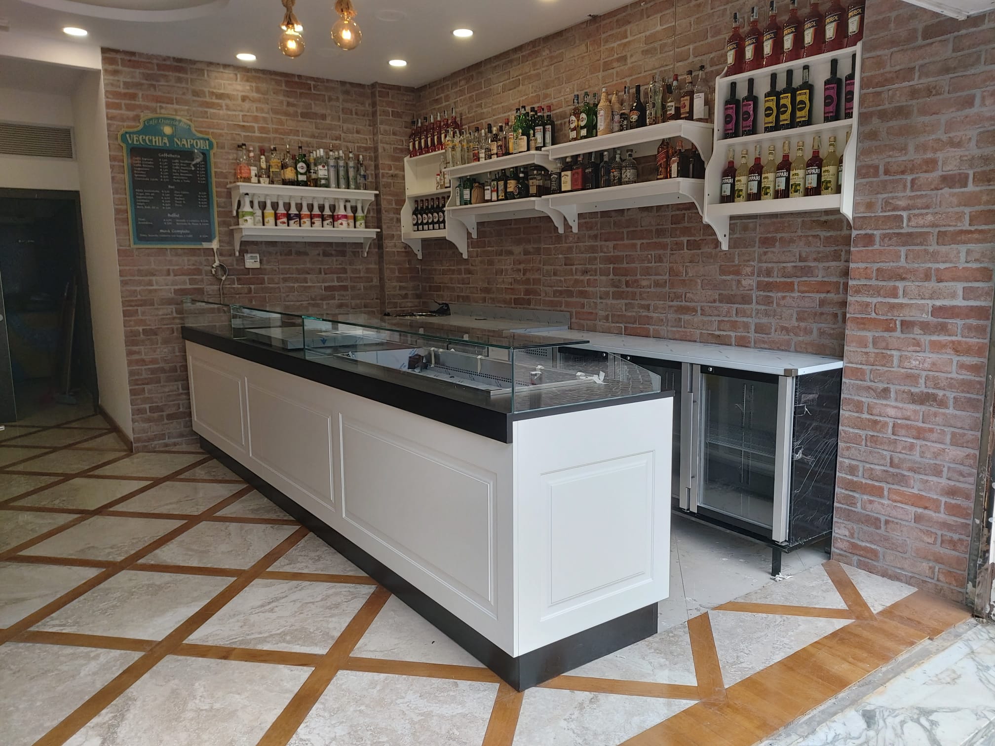 Interno di un bar moderno con un bancone in legno bianco, ripiani con bottiglie di liquori e una parete di mattoni a vista. Il pavimento è in marmo con intarsi in legno.
