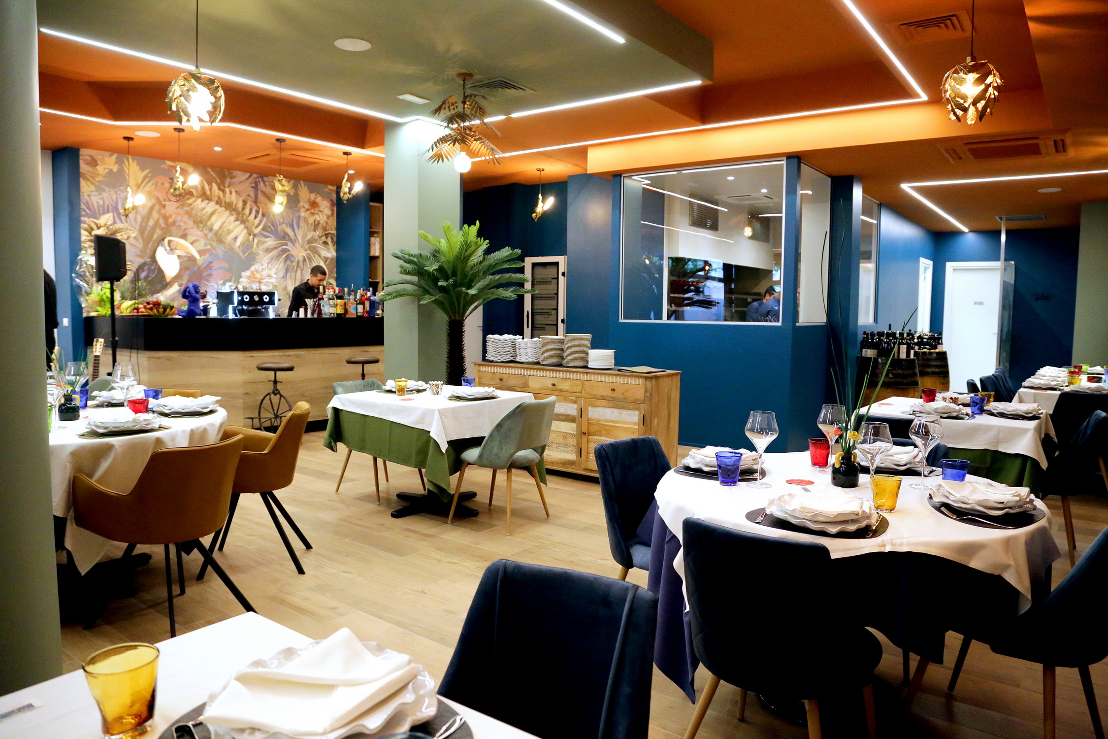 Interno di un ristorante a tema tropicale con un bancone bar, murale colorato, tavoli apparecchiati e decorazioni esotiche. Realizzato da Agape Arredo Bar.