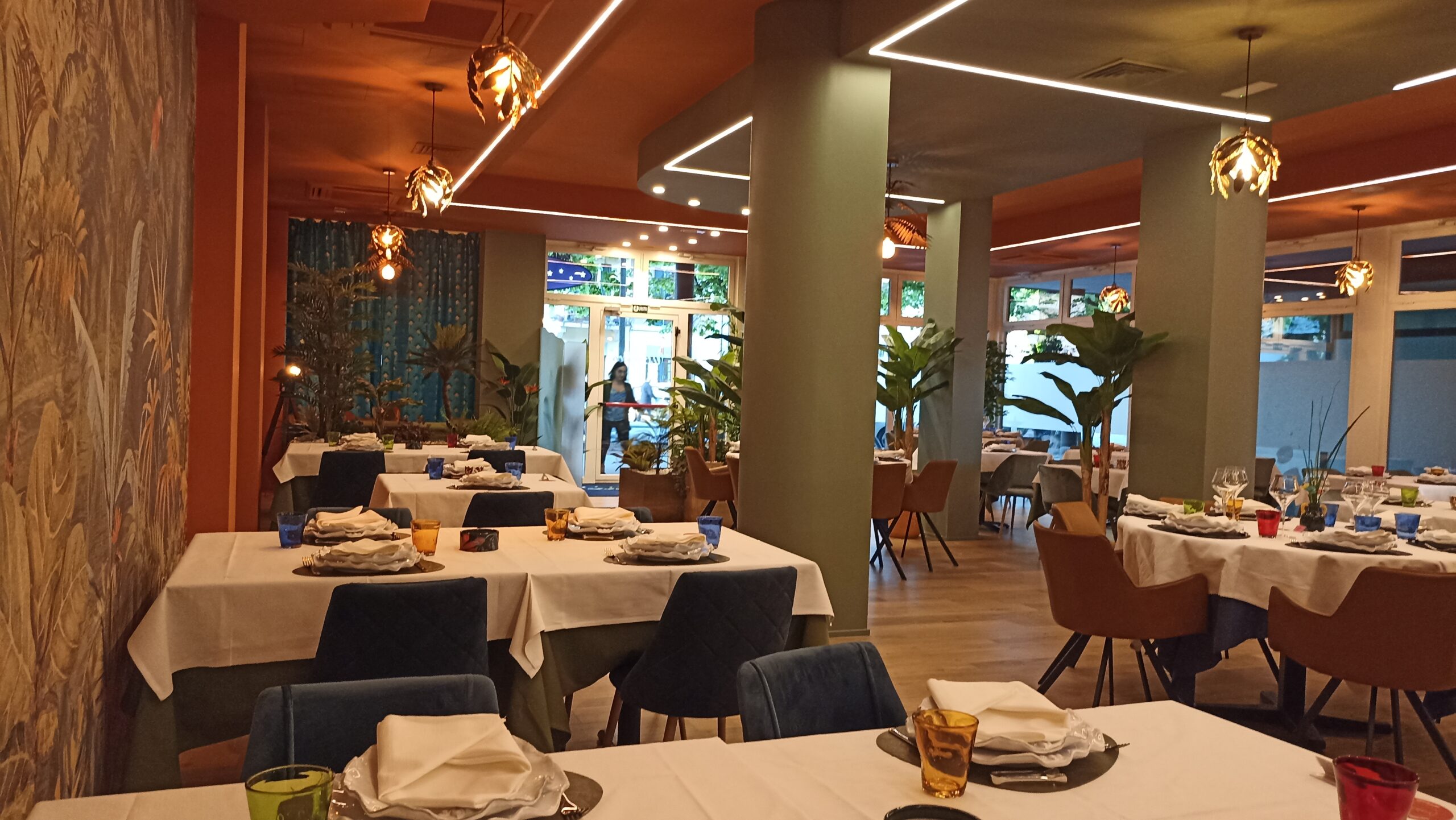 Interno di un ristorante elegante a tema tropicale con murale colorato, tavoli apparecchiati e illuminazione moderna. Realizzato da Agape Arredo Bar
