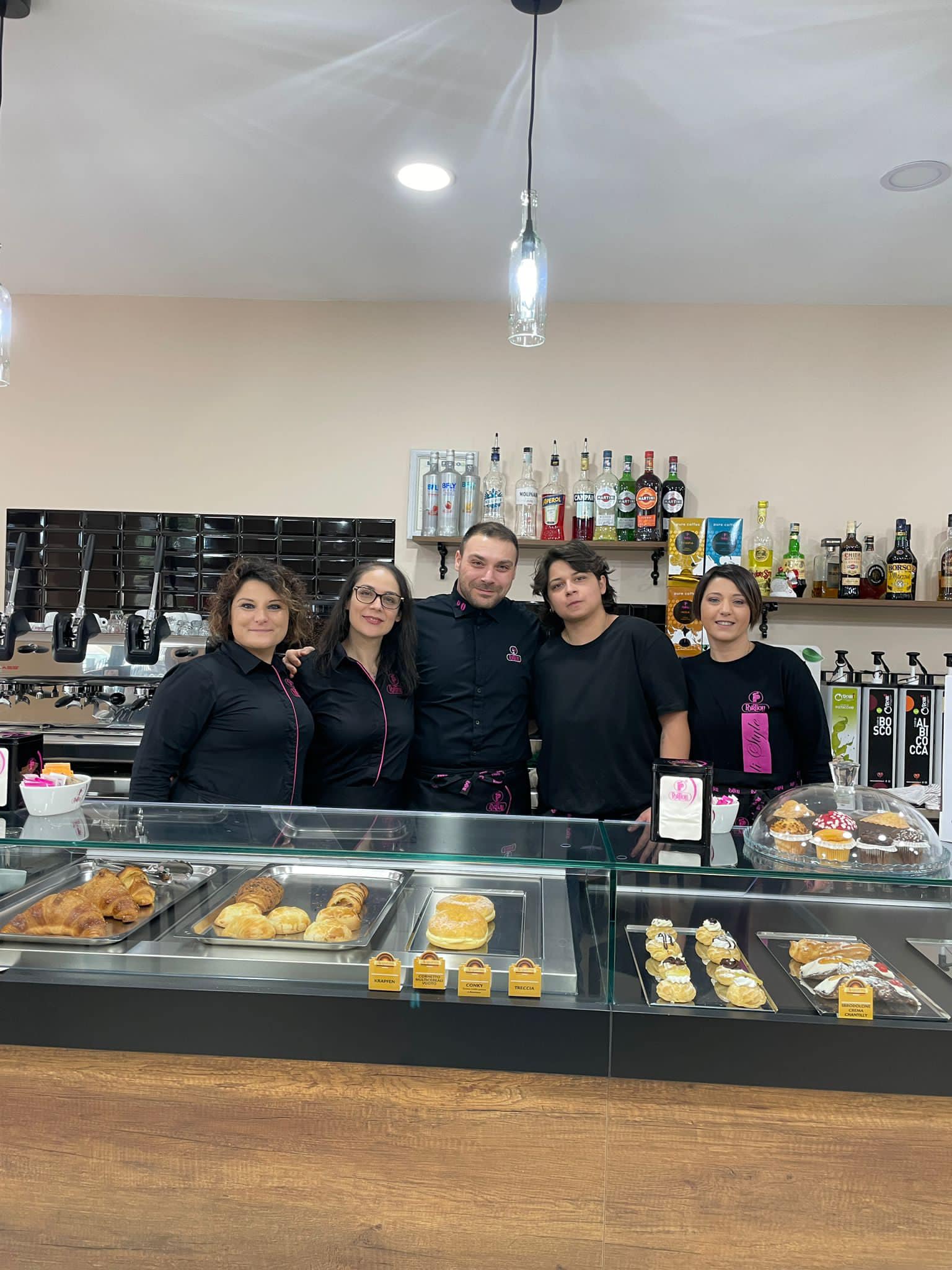 Staff di un bar moderno dietro al bancone con prodotti da forno e liquori, progettato da Agape Arredo Bar