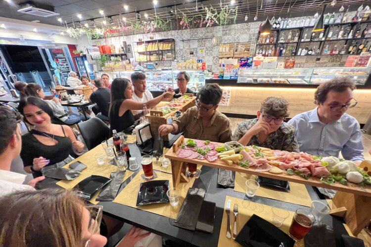 Gruppo di persone a tavola in un locale moderno, condividendo un lungo piatto di salumi e formaggi