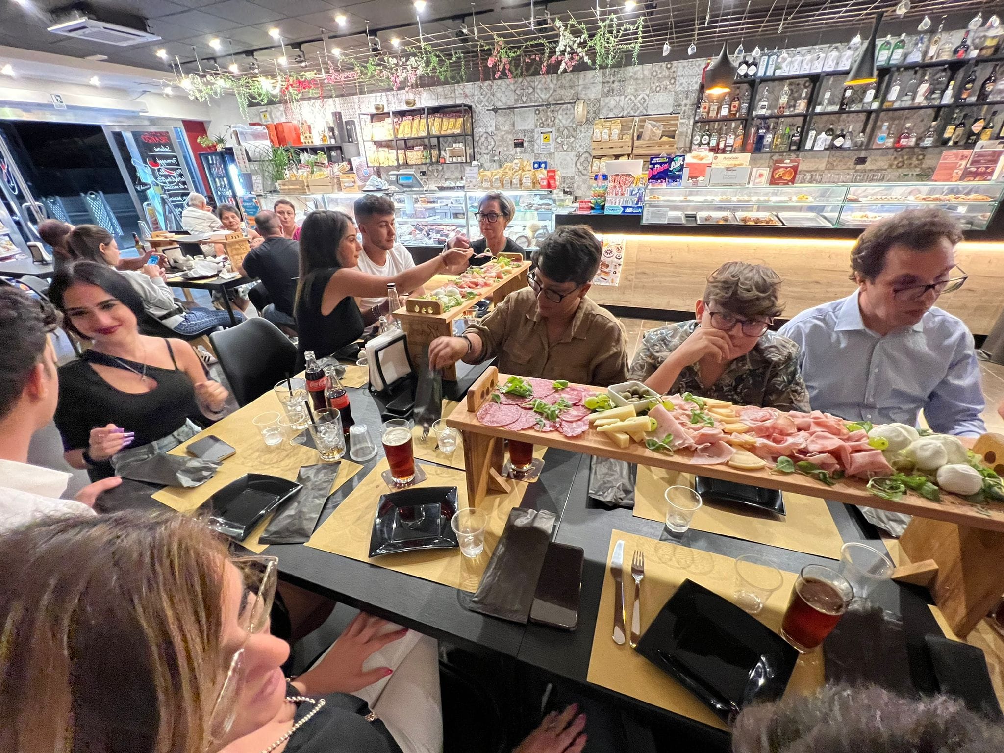 Gruppo di persone a tavola in un locale moderno, condividendo un lungo piatto di salumi e formaggi