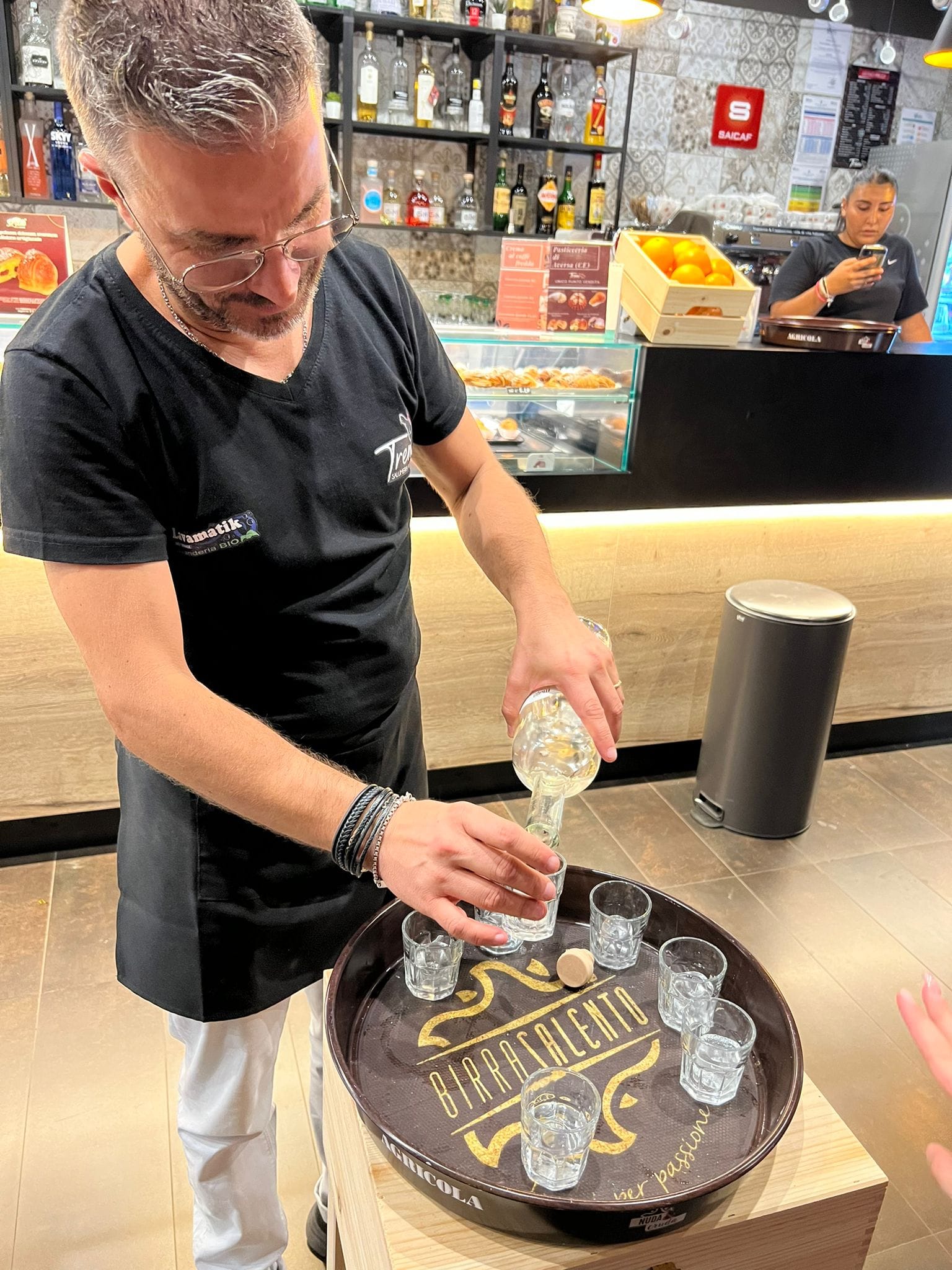 Barista che versa una bevanda in bicchieri su un vassoio, con scaffali di bottiglie sullo sfondo