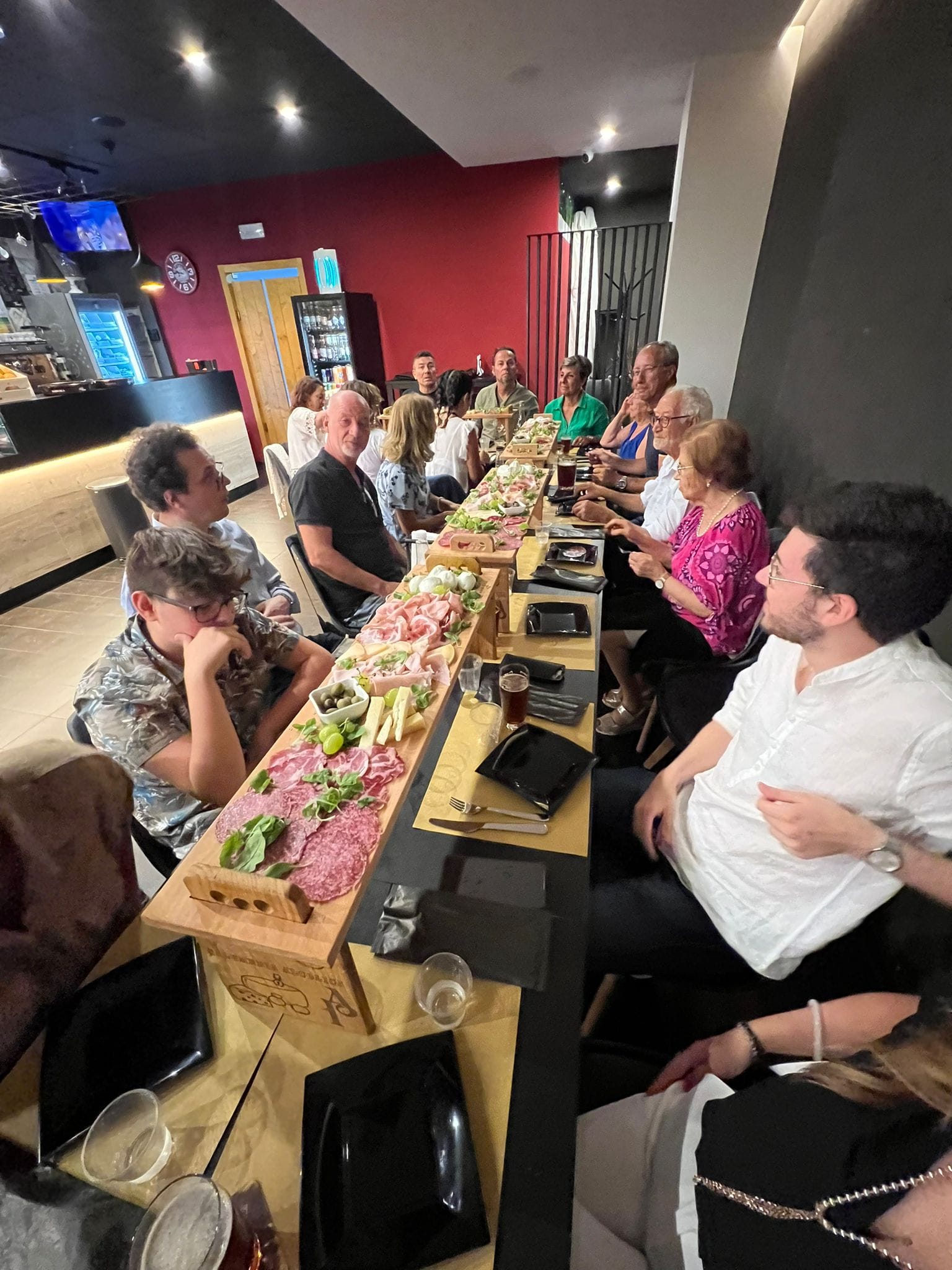 Gruppo di persone a tavola in un locale moderno, con un lungo piatto di salumi e formaggi
