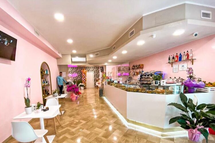 Locale con banco angolare bianco 6 metri, zoccolo dorato, vetrato sopra con colazioni e dolci, pareti rosa, pavimento parquet chiaro, sedie e tavoli bianchi, realizzato da Agape Arredo Bar
