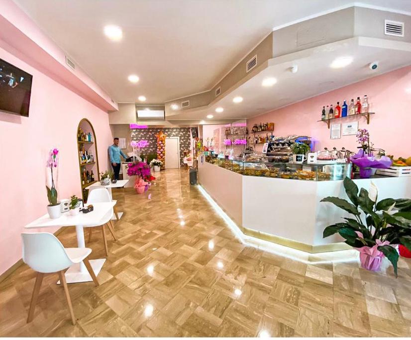 Locale con banco angolare bianco 6 metri, zoccolo dorato, vetrato sopra con colazioni e dolci, pareti rosa, pavimento parquet chiaro, sedie e tavoli bianchi, realizzato da Agape Arredo Bar