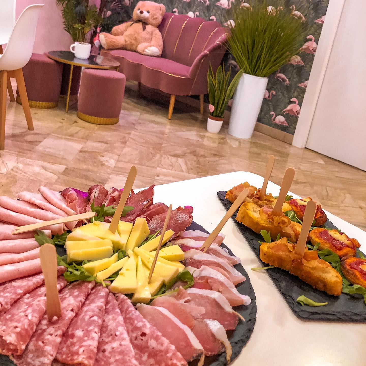 Buffet aperitivo con salumi e formaggi, divanetto e pouff area selfie con carta da parati fenicotteri rosa, realizzato da Agape Arredo Bar