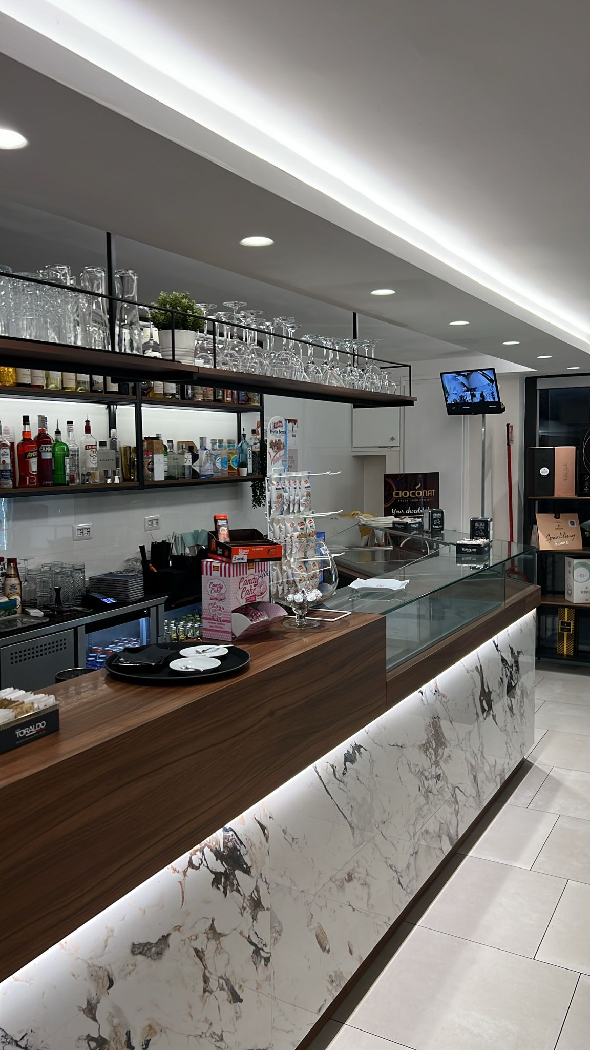 Interno di un locale con bancone bar moderno, ripiani in legno e marmo, scaffali con bicchieri e bottiglie